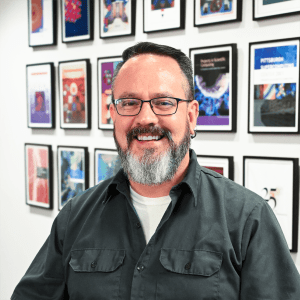 A man with light toned skin, brown hair, and a beard smiles at the camera.