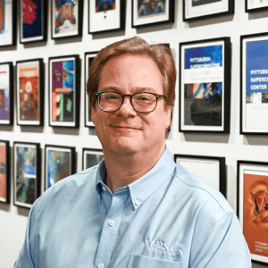 A man white light toned skin, blonde hair, and glasses smiles at the camera.