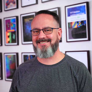 A man with light toned skin, brown hair, and a beard smiles at the camera.